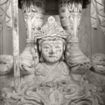 Black and white photograph by Bernard Donjean that shows the wooden sculpture of a Buddhist character carrying a divine guard on his hands and shoulders (name of the photograph : Strong Porter)