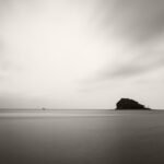 Black and white photograph by Bernard Donjean that shows a small boat sailing on a calm sea near an island situated in the Wakayama Prefecture (name of the photograph : The Island and the Boat)