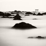 Black and white photograph by Bernard Donjean that shows a torii circeled by many rocks emerging from the sea (name of the photograph : Morito's Torii)